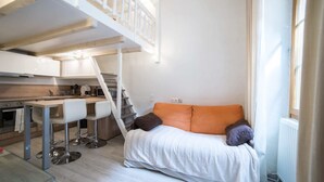 Un espace de vie compact comprenant un coin cuisine, un lit avec une literie aux couleurs orange et blanche, et un escalier menant à un niveau supérieur.