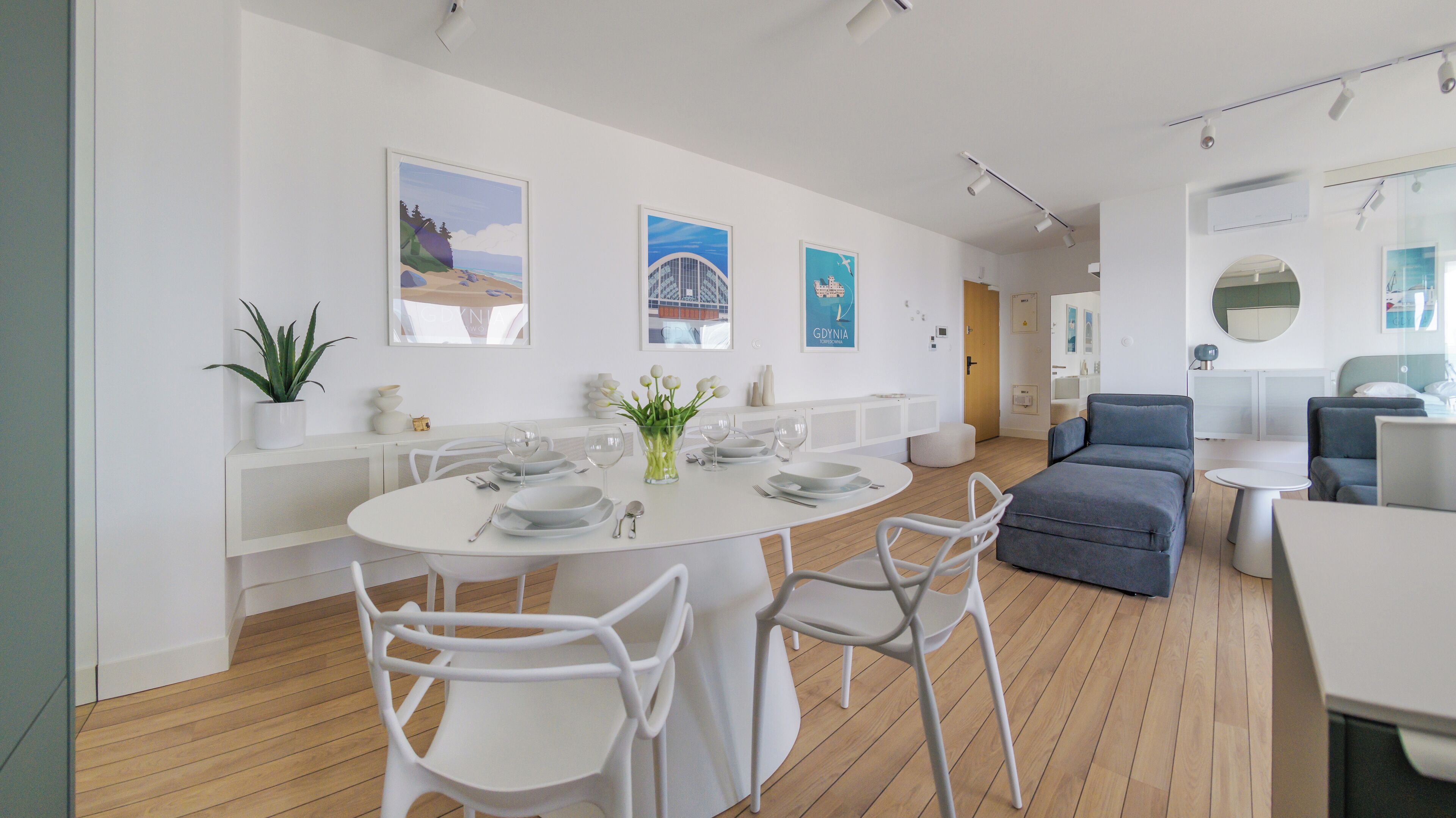 Une pièce moderne et bien éclairée, avec une table de salle à manger ronde préparée pour quatre personnes, des chaises blanches, un canapé gris et une petite table ronde.