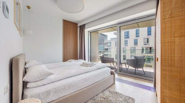 Une chambre moderne dotée d’un grand lit, offrant une vue sur la ville grâce à de larges fenêtres du sol au plafond, et d’un balcon agrémenté de meubles en osier.