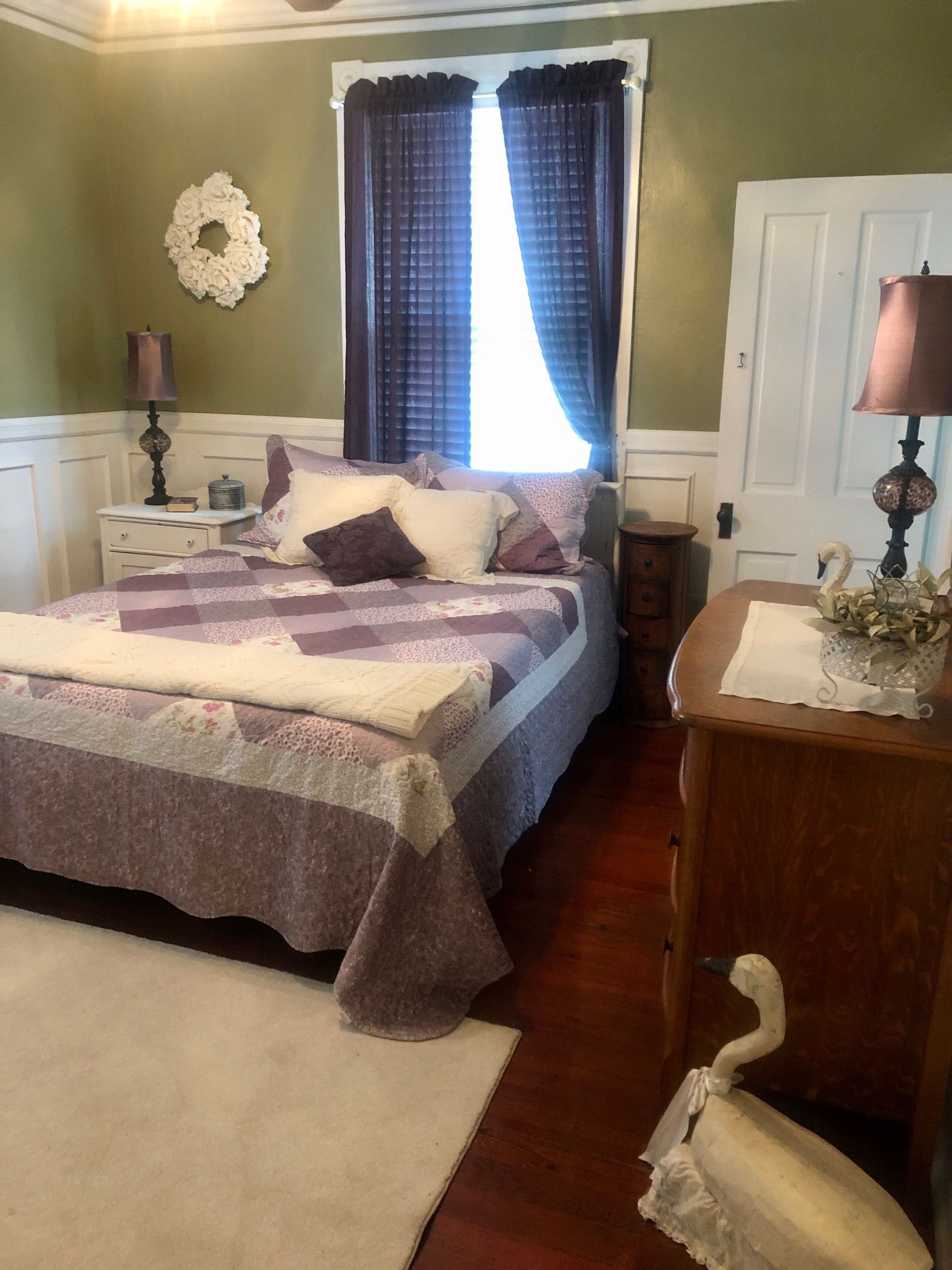 A bedroom with a bed, nightstands, a dresser, a lamp, a swan decoration, and a wreath on the wall.