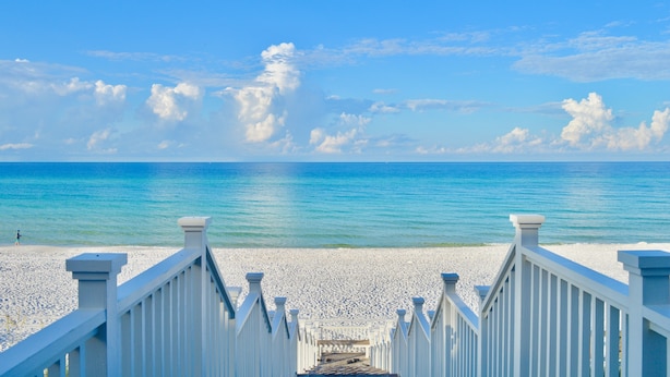 Une promenade en bois blanche qui mène à une plage, avec un ciel bleu dégagé et une mer calme.
