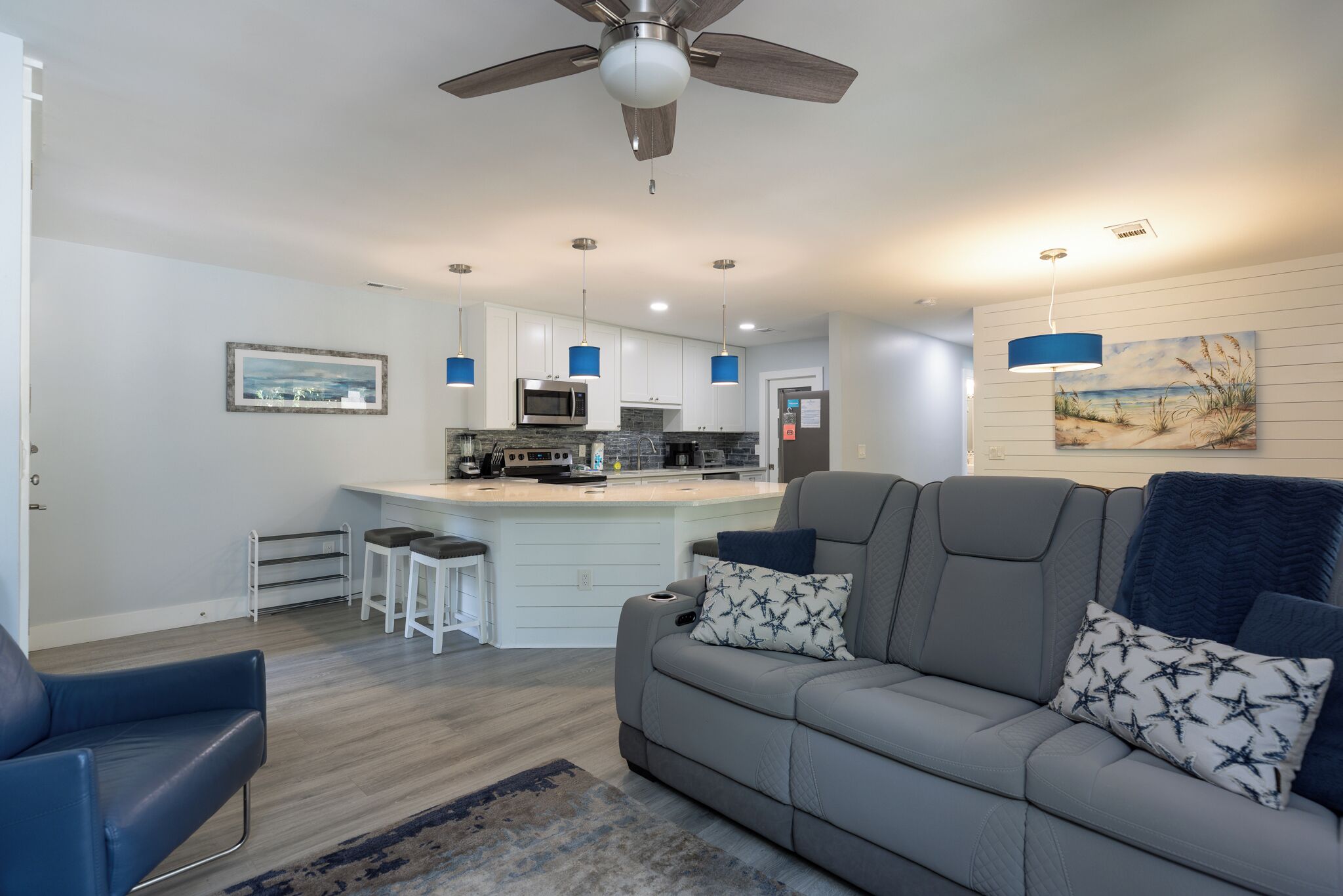 A modern living room with a grey sofa, a blue armchair, and a kitchen area with white cabinets and blue pendant lights.