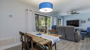 A modern living room with a dining area, a ceiling fan, a flat-screen TV, and a large window with curtains.