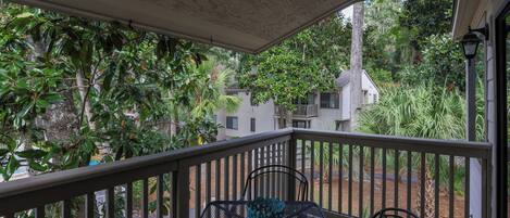 A covered balcony with a table and chairs, surrounded by lush greenery and residential buildings in the background.