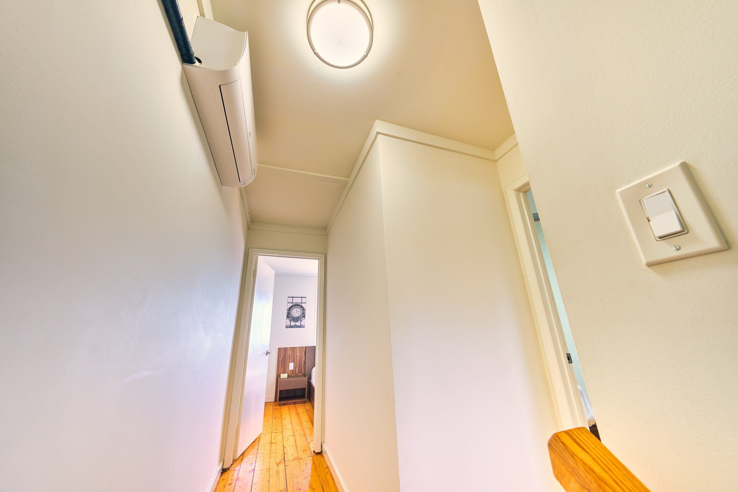 Un couloir étroit avec un plancher en bois, des murs blancs et un luminaire au plafond.