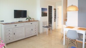 Ein modernes Wohnzimmer mit Flachbildfernseher, einem Essbereich mit Tisch und Stühlen sowie einer im Hintergrund sichtbaren Küche.
