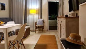 Ein gemütliches Wohnzimmer mit einer Couch, einem Esstisch, einem Sessel, einer Stehlampe, einem Fernseher und Blick ins Freie.