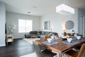 Un salon moderne avec un coin repas, comprenant une table à manger en bois pour six personnes, un canapé gris et une grande fenêtre laissant entrer la lumière naturelle.
