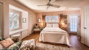 A bedroom with a large bed, a ceiling fan, a window with blinds, a chair, a nightstand, and a door. - Binky's Nest 1 Bedroom Home by RedAwning (Fredericksburg)