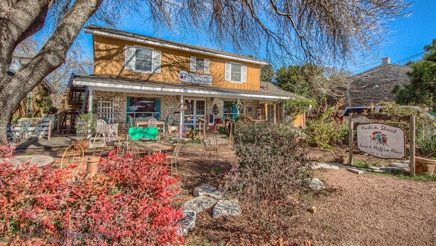 A two-story house with a front porch, a sign indicating 'Austin Street Java & Muffin Haus', and a garden area with red foliage and stone pathways. - Binky's Nest 1 Bedroom Home by RedAwning (Fredericksburg)