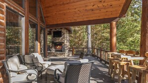 Una terraza de madera con mobiliario exterior, una chimenea de piedra y vistas del bosque circundante.