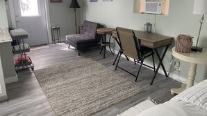 A room with a grey sofa, a small round table, a wooden desk, a chair, a rug, and a bed. There are framed pictures on the wall and a door with a sign. - Banana Beach Cottage (Lake Worth)