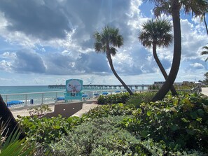 Une vue sur la plage avec un palmier, un panneau indiquant l’entrée de Lake Worth Beach, et une jetée en arrière-plan.