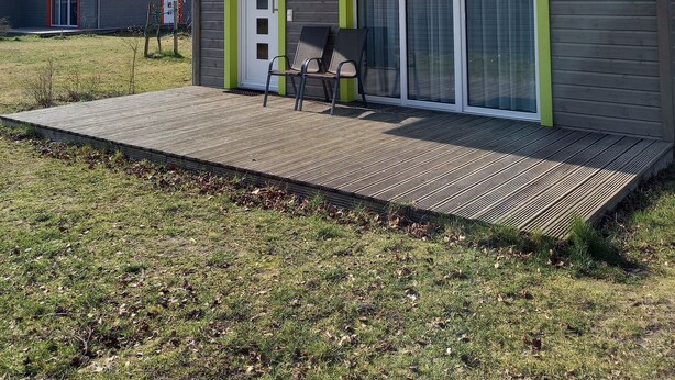 A small, single-story building with a green roof and a sliding glass door. There is a small deck area with two chairs.