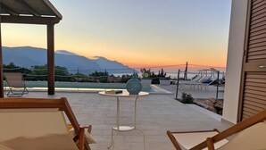 A patio with two wooden lounge chairs and a small table, overlooking a scenic view of mountains and a sunset.