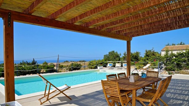 A covered outdoor area with a pool, wooden furniture, and a view of the sea.