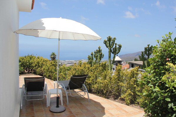 Un patio exterior con mesa y sillas bajo un paraguas blanco, con vistas a un paisaje de cactus y al mar.