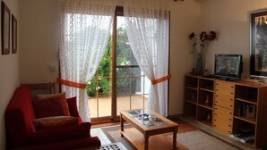 Ein Wohnzimmer mit einem roten Sofa, einem Glastisch, einem Fernseher auf einem Holzgestell und einer Glasschiebetür, die zu einem Garten führt.