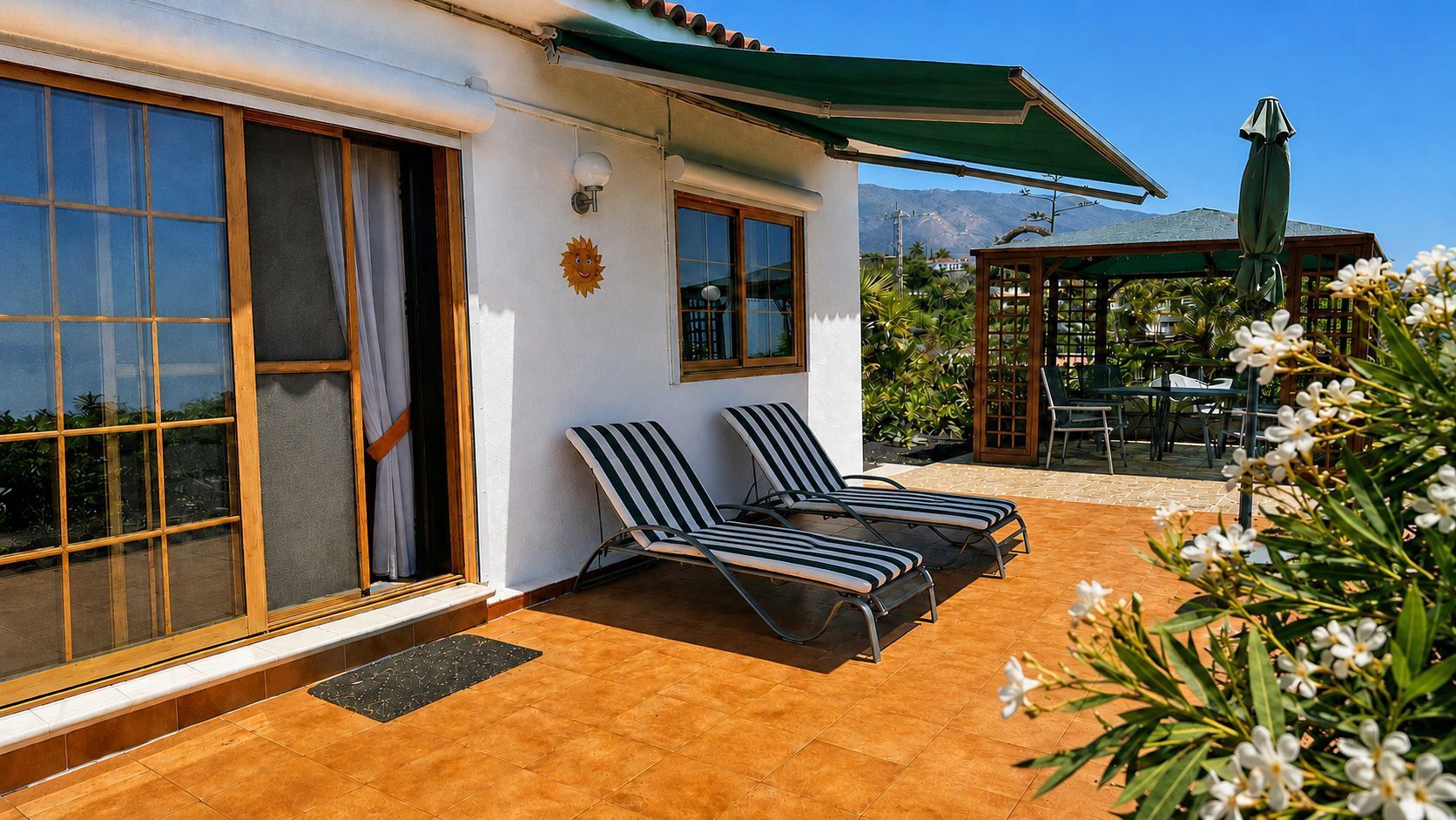 A two-story building with a patio area featuring two sun loungers, a covered seating area, and a view of mountains.