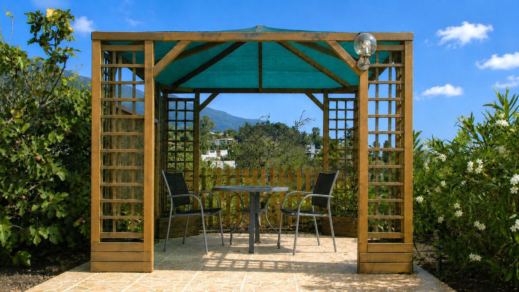 A wooden pergola with a table and chairs, surrounded by greenery and a mountain view.