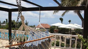 Een hangstoel, bevestigd aan een metalen frame, met uitzicht op een zwembadgedeelte met buitenmeubels en parasols.