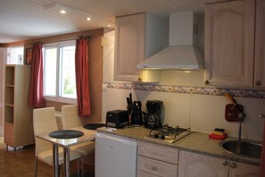 Eine kompakte Küche mit Gasherd, Kaffeemaschine und Toaster. Es gibt einen kleinen Kühlschrank, eine Spüle und ein Fenster mit Vorhängen.
