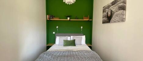 Een slaapkamer met een bed, groene muren, een hanglamp, een plank met boeken en een plant, en een lijst aan de muur.