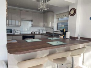 A modern kitchen with a central island, bar stools, a chandelier, and a clock on the wall.