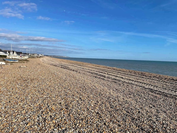 A pebbled beach with a clear sky, a few boats, and buildings in the background.