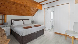 A bedroom with a wooden ceiling, a bed with pillows, a nightstand, a chair, and a door leading to another room. - Oceanfront Beach House - Family-Friendly (Pevensey)