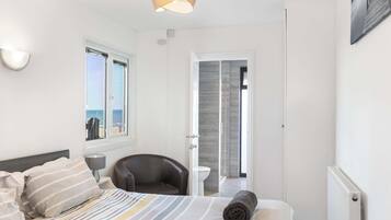Une chambre à coucher comprenant un lit, une chaise, une lampe, une fenêtre donnant sur la mer et une porte menant à une autre pièce.