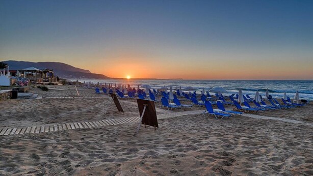 Una playa con filas de sillas azules y sombrillas, un paseo de madera y una puesta de sol al fondo.