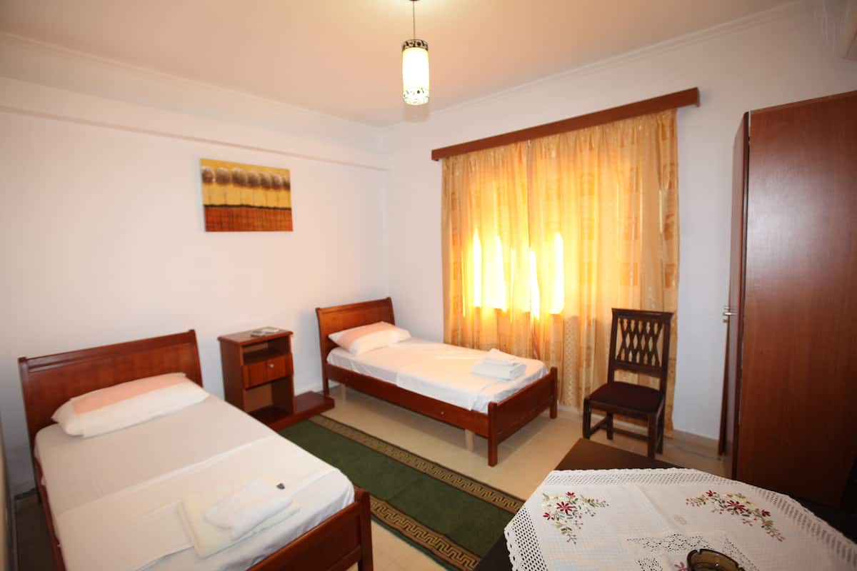 Quarto de hotel com duas camas de solteiro, uma cadeira de madeira, uma mesa pequena com toalha e uma porta.