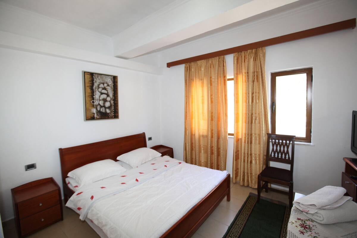 Quarto com cama de madeira, mesa de cabeceira, cadeira, televisão e janela com cortinas.