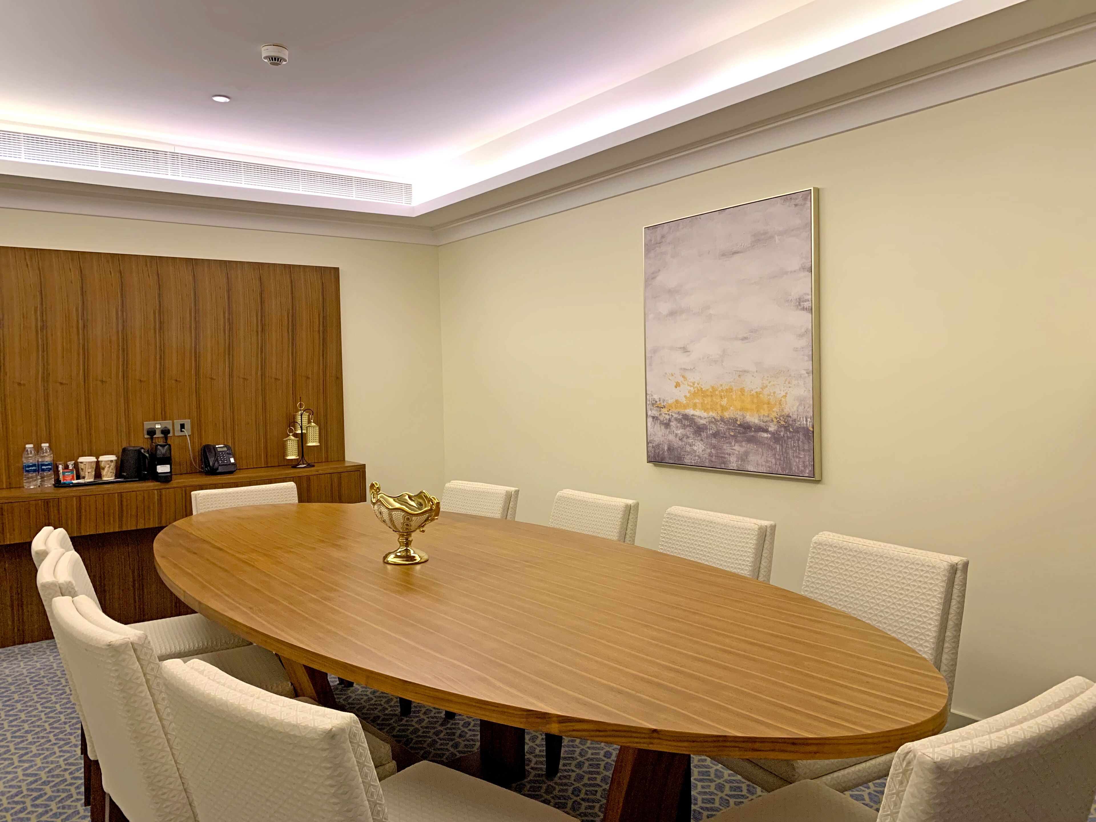 Un salón de conferencias con una mesa ovalada de madera, seis sillas tapizadas, un cuenco dorado decorativo, un cuadro abstracto colgado en la pared y un revestimiento de madera con un teléfono y una cafetera.