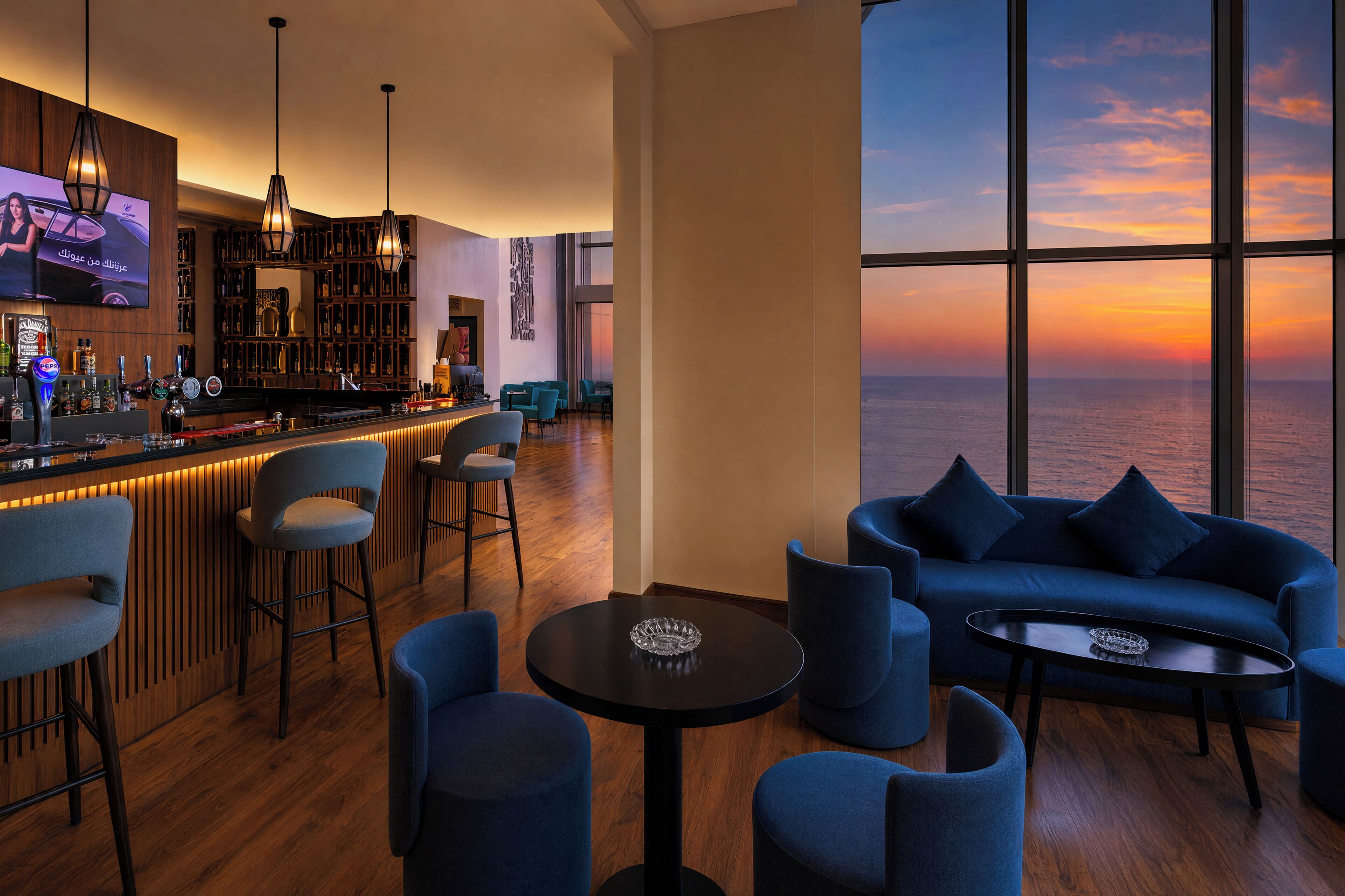 Zona de bar con taburetes, barra y una pantalla de televisión. Hay una sala de estar con sofás y sillones, y un amplio ventanal con vistas a la puesta de sol sobre el océano.