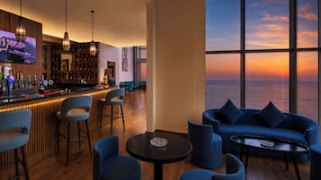 Zona de bar con taburetes, barra y una pantalla de televisión. Hay una sala de estar con sofás y sillones, y un amplio ventanal con vistas a la puesta de sol sobre el océano.