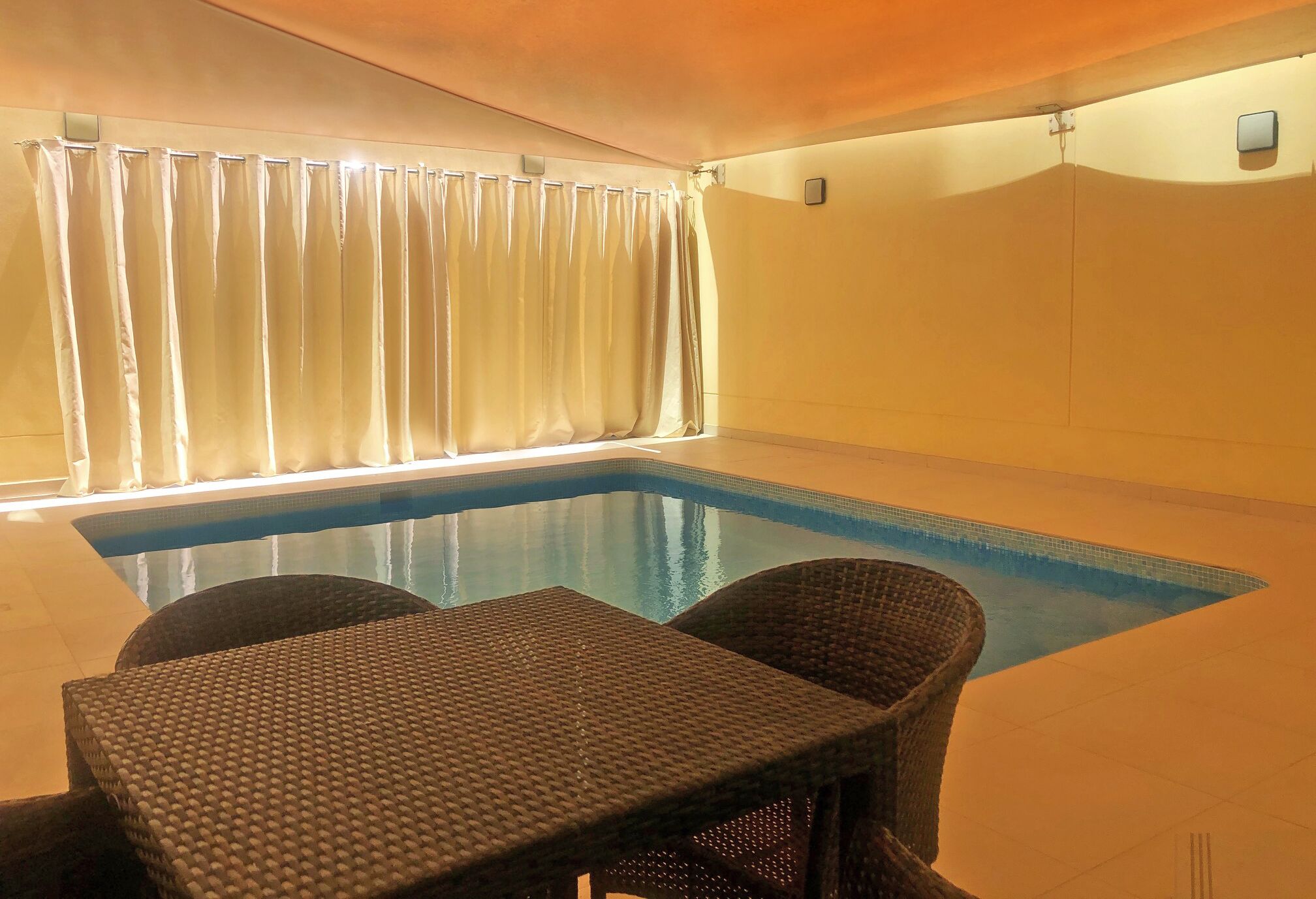 Una piscina interior con agua azul cristalina, rodeada de sillones de mimbre y una mesa.