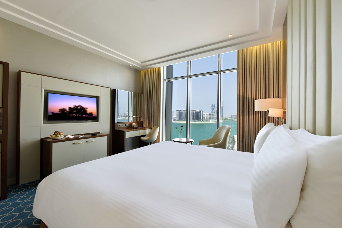 Habitación de hotel con una cama grande, un escritorio con televisor, una silla, una mesita y vistas a la ciudad.