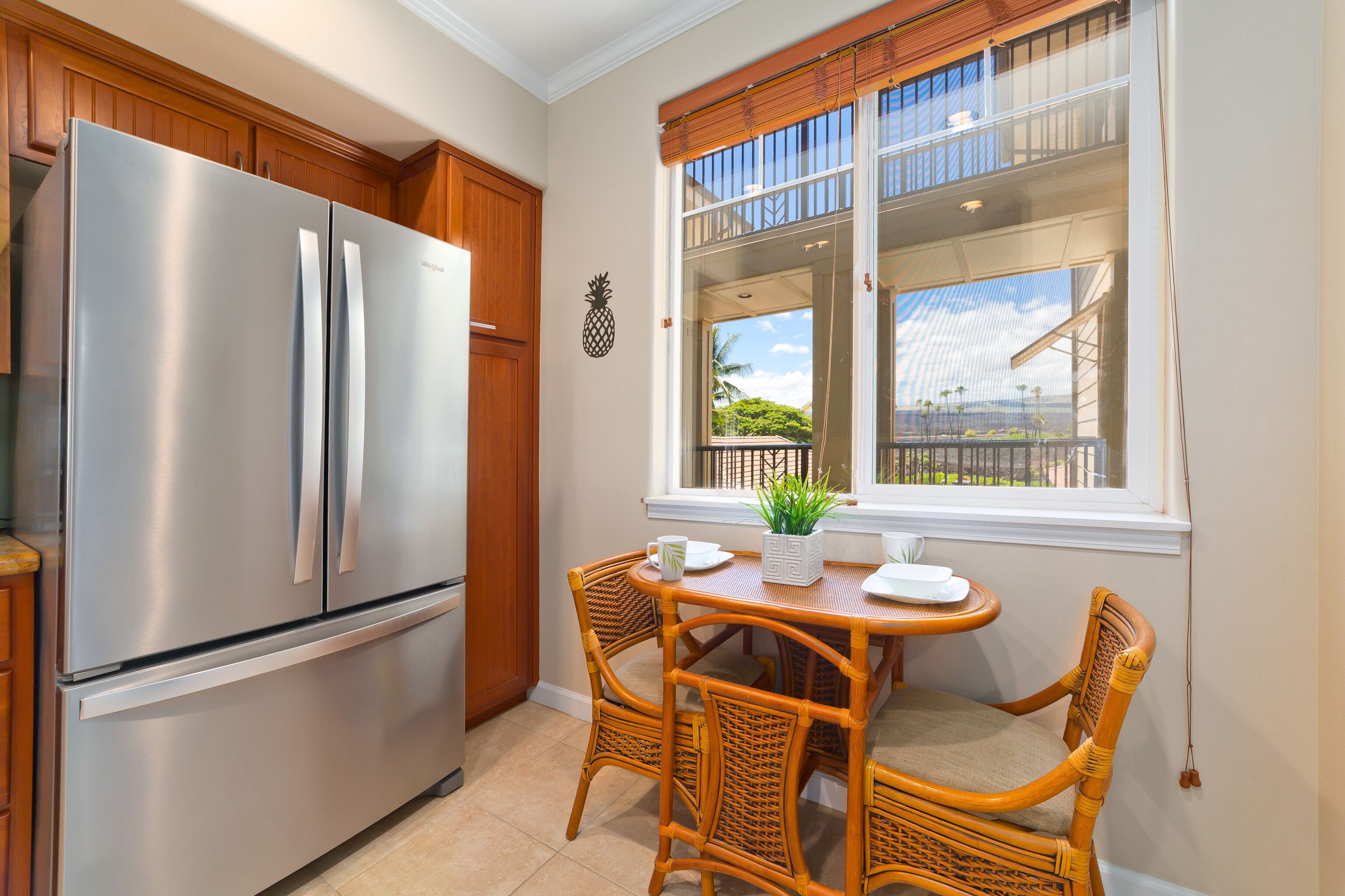 A kitchen with a stainless steel refrigerator, wooden cabinets, a dining table with wicker chairs, and a window with a view of greenery.