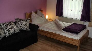 Une pièce comprenant un lit, un canapé noir avec des coussins à motifs, une fenêtre avec des rideaux et une table de chevet avec une lampe.