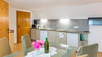 A modern kitchen with a glass dining table, chairs, a wooden door, and a flat-screen TV mounted on the wall.