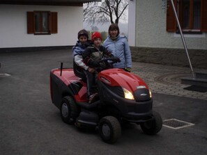 Drei Personen fahren mit einem roten Rasenmäher durch einen Innenhof, im Hintergrund ist ein Gebäude zu sehen.