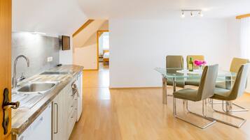 A modern kitchen with a glass dining table and chairs, a TV mounted on the wall, and a wooden floor.