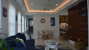 A modern living room with a blue sofa, a dining area with wicker chairs, a kitchen with wooden cabinets, and a ceiling fan. - Sweet Caroline Historic Seaport Key West Home Rental Floor 1 (Key West)