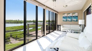 A spacious balcony with large windows overlooking a water body and greenery, a white sofa, and a ceiling fan. - Blue Lagoon Estate (Fort Myers Beach)