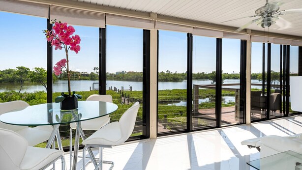 A modern room with large windows, a glass table, white chairs, and a view of a lake. - Blue Lagoon Estate (Fort Myers Beach)
