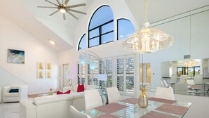 A spacious living room with a glass dining table, white sofas, and a large window with shutters. - Blue Lagoon Estate (Fort Myers Beach)
