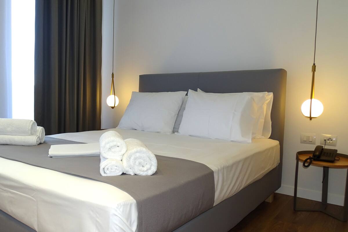A neatly made bed with white linens and rolled towels, a bedside phone, and a lamp on a nightstand.