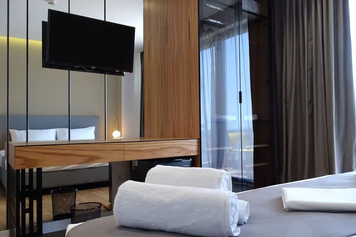 A hotel room with a bed, a TV mounted on the wall, a wooden headboard, a bedside table, and a window with curtains.